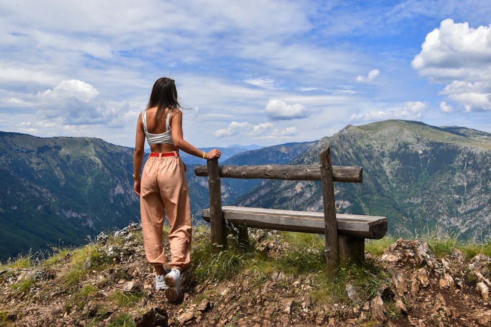 woman standing near wooden bench viewing mountain under blue and white skies during daytime