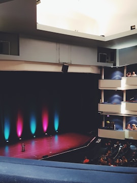 An empty theater stage is lit with colorful lights, including purple, blue, and pink hues. The stage has a single microphone stand and stool. The seating area is partially filled with an audience. The architectural design includes multiple balconies with blue walls and white railings. The lighting creates a vibrant and inviting atmosphere.
