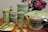 A collection of ceramic dinnerware designed to resemble asparagus, including a teapot, cups, and various containers. The set is adorned with green and pink hues that mimic the vegetable's natural appearance. In the background, a bouquet of pink flowers adds a vibrant touch.