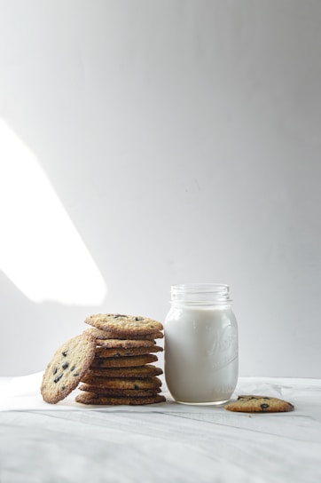 A stack of chocolate chip cookies is placed next to a mason jar filled with milk. One cookie lies separately in front of the jar. The scene is set against a simple, light-colored background, with soft lighting creating a cozy and inviting atmosphere.