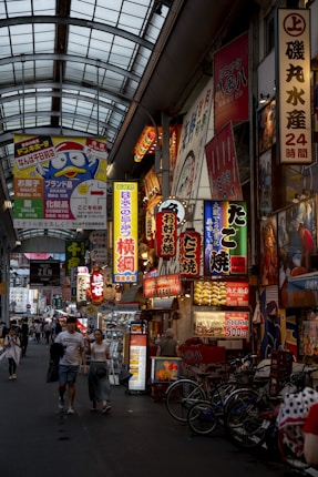 A bustling urban street market filled with vibrant neon signs advertising various shops and products. People walk through the narrow pathway between the shops. The market is covered by a metal and glass roof, which adds to the enclosed feel of the area. Numerous bicycles are parked to the side, and a mix of Japanese text dominates the signage.