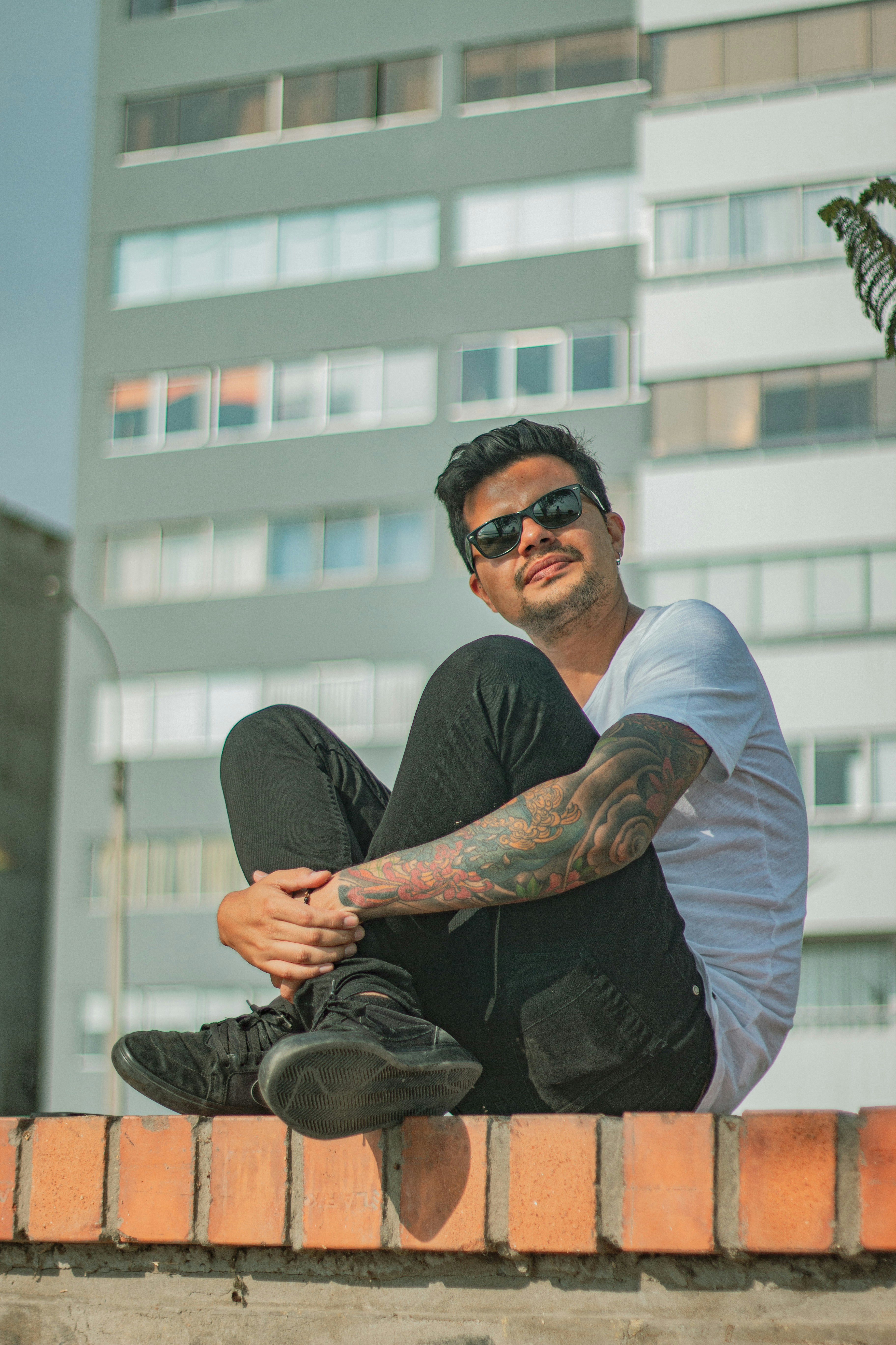 man wearing white shirt and black denim jeans sitting on concrete surface near building during daytime