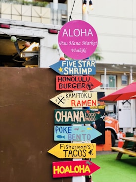 Colorful directional signs are displayed on a pole, each pointing to different food options such as shrimp, burger, ramen, loco moco, bento, and tacos. The backdrop features a food truck and a picnic area with a green surface and red umbrellas.