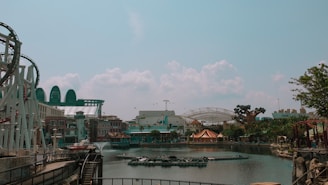 An amusement park scene featuring a roller coaster on the left, surrounded by water and various themed buildings. In the background, there are structures resembling futuristic domes and a large artificial tree. The sky is clear with a few clouds, lending a calm atmosphere to the park.