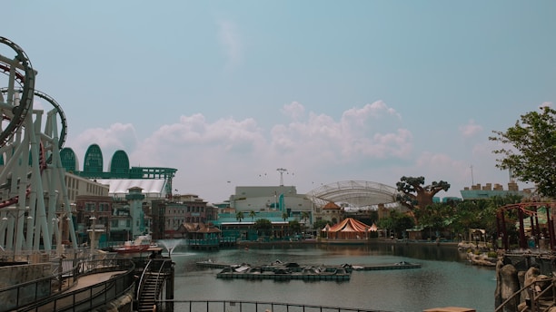 An amusement park scene featuring a roller coaster on the left, surrounded by water and various themed buildings. In the background, there are structures resembling futuristic domes and a large artificial tree. The sky is clear with a few clouds, lending a calm atmosphere to the park.