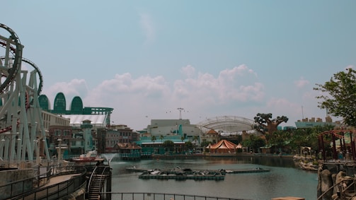 An amusement park scene featuring a roller coaster on the left, surrounded by water and various themed buildings. In the background, there are structures resembling futuristic domes and a large artificial tree. The sky is clear with a few clouds, lending a calm atmosphere to the park.