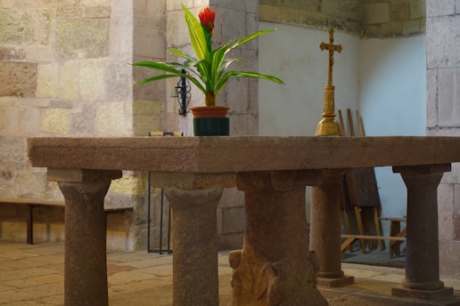 A sturdy stone table with four thick columns stands in a stone-walled room. On the table, there is a small red and green potted plant alongside a golden religious cross. The background includes a few wooden items leaning against the wall.