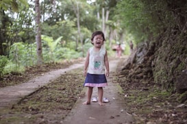 A young child stands on a moss-covered pathway in a lush, green outdoor setting. The child appears to be crying or upset, wearing a sleeveless top with a cartoon character and a blue skirt with pink trim. The surrounding environment is rich with vegetation, including trees and bushes along the path.