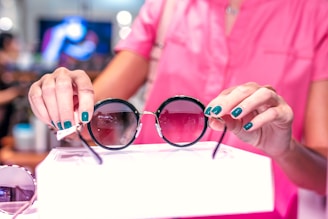A person with teal-colored nails is holding a pair of stylish round sunglasses. The individual is wearing a pink top, and the background is blurred with bokeh lights, suggesting a retail or shopping environment.