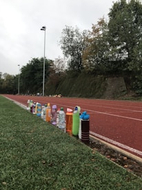 A row of colorful water bottles lined up on a wet trackside, with a running track and lush green foliage in the background. The sky appears overcast, creating a slightly moody atmosphere.