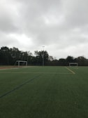 An empty soccer field with green artificial turf is depicted. Two goalposts are positioned on opposite sides of the field. The sky is overcast with thick grey clouds, and trees surround the field, indicating it is located outdoors.