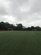 An empty soccer field with green artificial turf is depicted. Two goalposts are positioned on opposite sides of the field. The sky is overcast with thick grey clouds, and trees surround the field, indicating it is located outdoors.