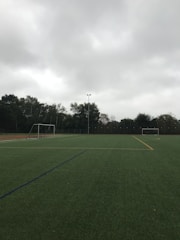 An empty soccer field with green artificial turf is depicted. Two goalposts are positioned on opposite sides of the field. The sky is overcast with thick grey clouds, and trees surround the field, indicating it is located outdoors.