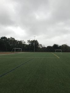An empty soccer field with green artificial turf is depicted. Two goalposts are positioned on opposite sides of the field. The sky is overcast with thick grey clouds, and trees surround the field, indicating it is located outdoors.