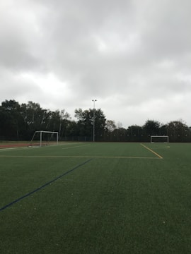 An empty soccer field with green artificial turf is depicted. Two goalposts are positioned on opposite sides of the field. The sky is overcast with thick grey clouds, and trees surround the field, indicating it is located outdoors.