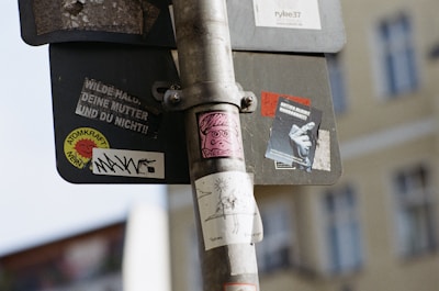 A street sign covered with various stickers, featuring designs and text in different languages. One sticker depicts a pink cartoon face, while another shows a black and white photograph of a person. The pole has multiple layers of adhesive labels, creating a layered and textured look. The background showcases a blurred view of a yellow building.