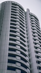 A modern high-rise building with a distinctive curved design featuring multiple balconies. The architecture is sleek and predominantly composed of white and gray tones, conveying a sense of minimalism and contemporary style.