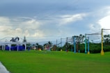 A grassy outdoor space featuring a row of colorful posts supporting rope nets, which appear to be part of an obstacle or climbing course. To the left, there is a white-frame shelter with a stack of blue chairs underneath it. The sky is overcast with gray and white clouds, giving the scene a calm appearance. Trees and bushes can be seen in the background.