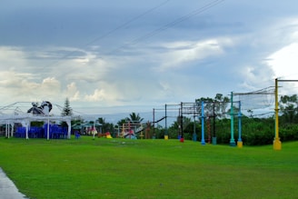 A grassy outdoor space featuring a row of colorful posts supporting rope nets, which appear to be part of an obstacle or climbing course. To the left, there is a white-frame shelter with a stack of blue chairs underneath it. The sky is overcast with gray and white clouds, giving the scene a calm appearance. Trees and bushes can be seen in the background.