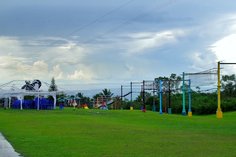 A grassy outdoor space featuring a row of colorful posts supporting rope nets, which appear to be part of an obstacle or climbing course. To the left, there is a white-frame shelter with a stack of blue chairs underneath it. The sky is overcast with gray and white clouds, giving the scene a calm appearance. Trees and bushes can be seen in the background.
