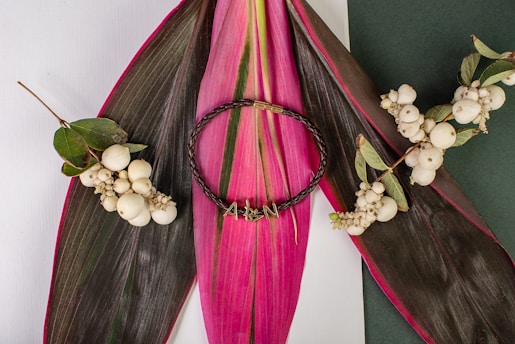 A braided dark brown bracelet with gold accents is placed on large, vibrant pink and green tropical leaves. The bracelet is surrounded by clusters of white berries attached to green stems, creating a natural and artistic arrangement.