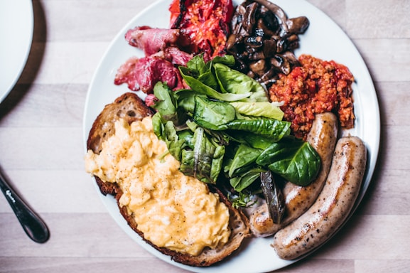 An appetizing breakfast plate featuring scrambled eggs on toast, fresh greens, sausages, sautéed mushrooms, roasted tomatoes, crispy bacon, and a serving of savory minced meat.