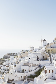A picturesque view of a coastal town with iconic whitewashed buildings and blue-domed roofs, perched on a hillside. Windmills are visible in the background, and a few people are scattered throughout the scene. The sky is clear, suggesting a sunny day, and the buildings cascade down towards the sea.