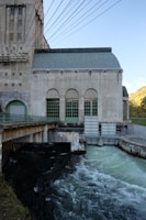 A large industrial building with arched windows, displaying a concrete structure. Numerous cables span overhead, suggesting it might be a power plant. Below, water flows rapidly, indicating the presence of a dam or hydroelectric facility. The backdrop includes a clear sky and distant hills.