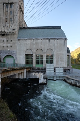 A large industrial building with arched windows, displaying a concrete structure. Numerous cables span overhead, suggesting it might be a power plant. Below, water flows rapidly, indicating the presence of a dam or hydroelectric facility. The backdrop includes a clear sky and distant hills.