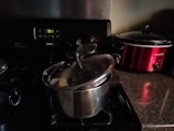 A kitchen scene featuring a stove with a stainless steel pot and lid. To the right, there is a red Crock-Pot on a granite countertop. The stove's display shows the time as 7:01.