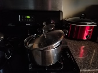 A kitchen scene featuring a stove with a stainless steel pot and lid. To the right, there is a red Crock-Pot on a granite countertop. The stove's display shows the time as 7:01.