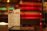 A menu card stands upright on a table in a dimly lit setting, with a colorful blurred background of red and golden hues. The text on the menu is in Russian, and other items like a small device or holder and papers are visible around it.