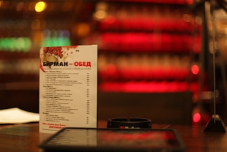 A menu card stands upright on a table in a dimly lit setting, with a colorful blurred background of red and golden hues. The text on the menu is in Russian, and other items like a small device or holder and papers are visible around it.