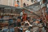 A person sits reading a book amidst an eclectic outdoor stairway made entirely of colorful, stacked books. The surrounding walls are rustic brick, adorned with phrases encouraging exploration. A decorative ship's wheel is visible, adding a nautical theme.