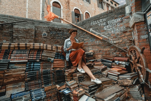 A person sits reading a book amidst an eclectic outdoor stairway made entirely of colorful, stacked books. The surrounding walls are rustic brick, adorned with phrases encouraging exploration. A decorative ship's wheel is visible, adding a nautical theme.