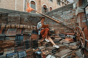 A person sits reading a book amidst an eclectic outdoor stairway made entirely of colorful, stacked books. The surrounding walls are rustic brick, adorned with phrases encouraging exploration. A decorative ship's wheel is visible, adding a nautical theme.