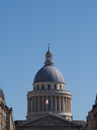 A neoclassical dome structure dominates the scene, resembling the architecture of a grand historical building. The building appears to have a French flag hanging from its facade, emphasizing its national significance. The sky is clear and blue, providing a striking backdrop.