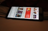 A smartphone displaying a streaming app interface, showing various movie and TV show thumbnails. The screen includes categories like 'For you' and 'New release hit films', with images from popular films. The background is a dimly lit setting with a fabric surface.
