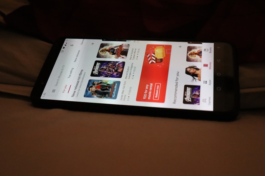 A smartphone displaying a streaming app interface, showing various movie and TV show thumbnails. The screen includes categories like 'For you' and 'New release hit films', with images from popular films. The background is a dimly lit setting with a fabric surface.