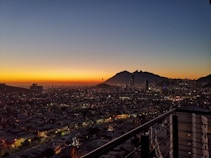 A vibrant cityscape at dusk with a glowing orange horizon transitioning to a deep blue sky. High-rise buildings and a mountain silhouette dominate the skyline, while city lights begin to sparkle across the densely packed urban area below. A balcony with metal railings is visible in the foreground.