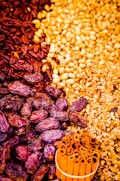 An assortment of dried fruits and nuts carefully arranged together. Dark red dates on the left merge into a cluster of yellow grains or seeds, transitioning to pieces of cracked walnuts. At the bottom, there is a bundle of cinnamon sticks tied together.