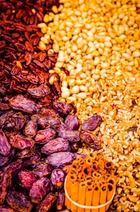 An assortment of dried fruits and nuts carefully arranged together. Dark red dates on the left merge into a cluster of yellow grains or seeds, transitioning to pieces of cracked walnuts. At the bottom, there is a bundle of cinnamon sticks tied together.
