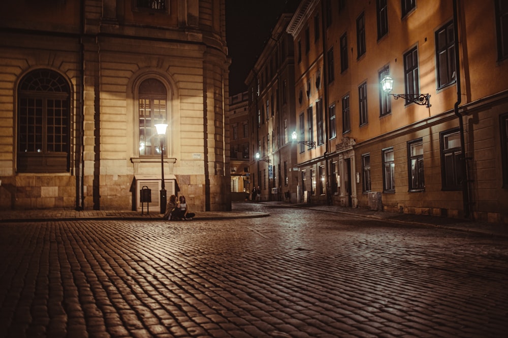 two persons sitting on curb beside lamppost near buildings during night