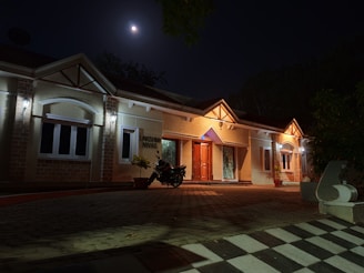 A single-story house illuminated by exterior lights at night. The moon shines brightly in the sky above, casting a soft light over the scene. A motorcycle is parked near the entrance, and a checkered pattern walkway leads to the house. Plants in pots are placed near the entrance door.