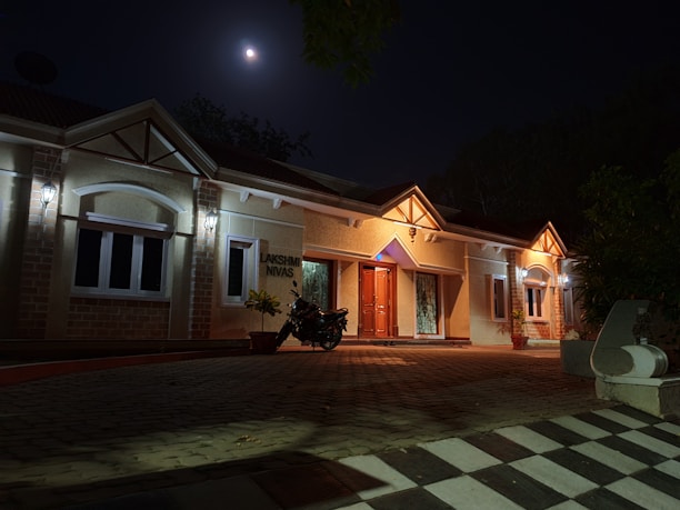 A single-story house illuminated by exterior lights at night. The moon shines brightly in the sky above, casting a soft light over the scene. A motorcycle is parked near the entrance, and a checkered pattern walkway leads to the house. Plants in pots are placed near the entrance door.
