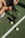 A person sits on a tennis court, holding a tennis racket with a green and black color scheme. Two tennis balls and a can of tennis balls are positioned near the racket. The person is wearing white athletic shoes and dark sportswear. The court is a typical green tennis surface, and a net is visible nearby.