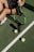 A person sits on a tennis court, holding a tennis racket with a green and black color scheme. Two tennis balls and a can of tennis balls are positioned near the racket. The person is wearing white athletic shoes and dark sportswear. The court is a typical green tennis surface, and a net is visible nearby.