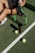 A person sits on a tennis court, holding a tennis racket with a green and black color scheme. Two tennis balls and a can of tennis balls are positioned near the racket. The person is wearing white athletic shoes and dark sportswear. The court is a typical green tennis surface, and a net is visible nearby.
