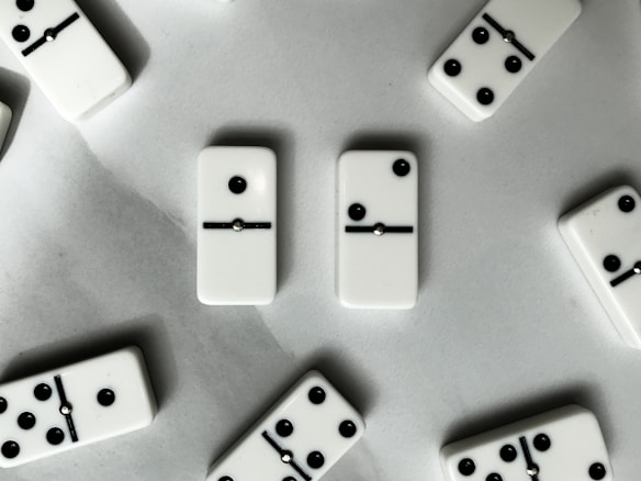 A collection of domino tiles scattered on a light-colored surface. The tiles display a variety of black dots in different configurations with a black line dividing the two halves. The arrangement appears random, with some tiles showing a single dot and others revealing multiple dots up to five.