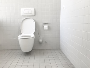 A clean and minimalistic bathroom featuring a wall-mounted toilet with an open lid. The walls and floor are tiled in a light gray color. A roll of toilet paper is mounted on the wall nearby, accompanied by a toilet brush in a holder.
